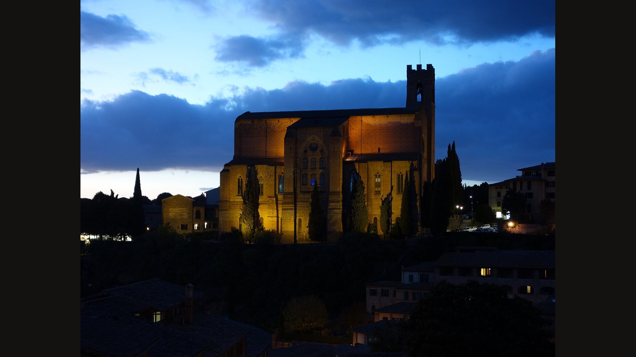 The Basilica of San Domenico in Siena at dusk, its brick walls and square bell tower lit warm gold against a deep blue cloudy sky, with dark cypress trees beside it