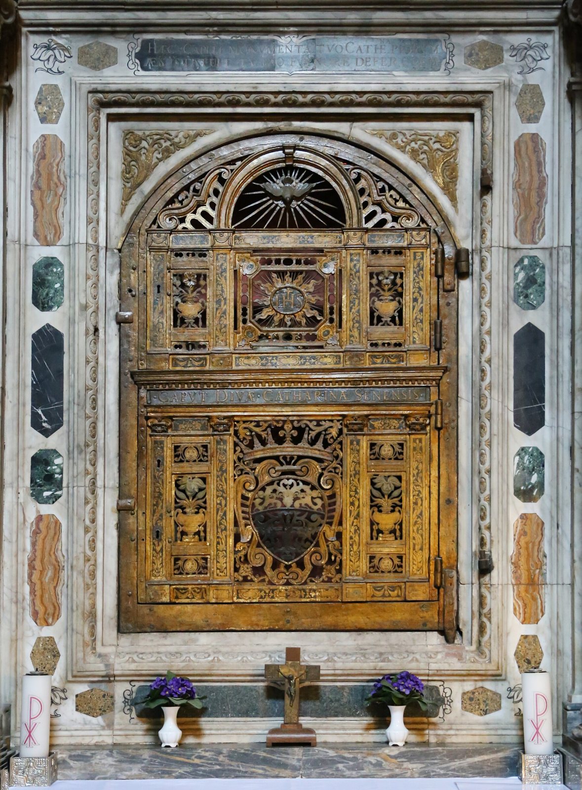 A tall marble altar frame in San Domenico holding a closed, ornate gilded grille and door, with a small crucifix and two vases of violets at its base