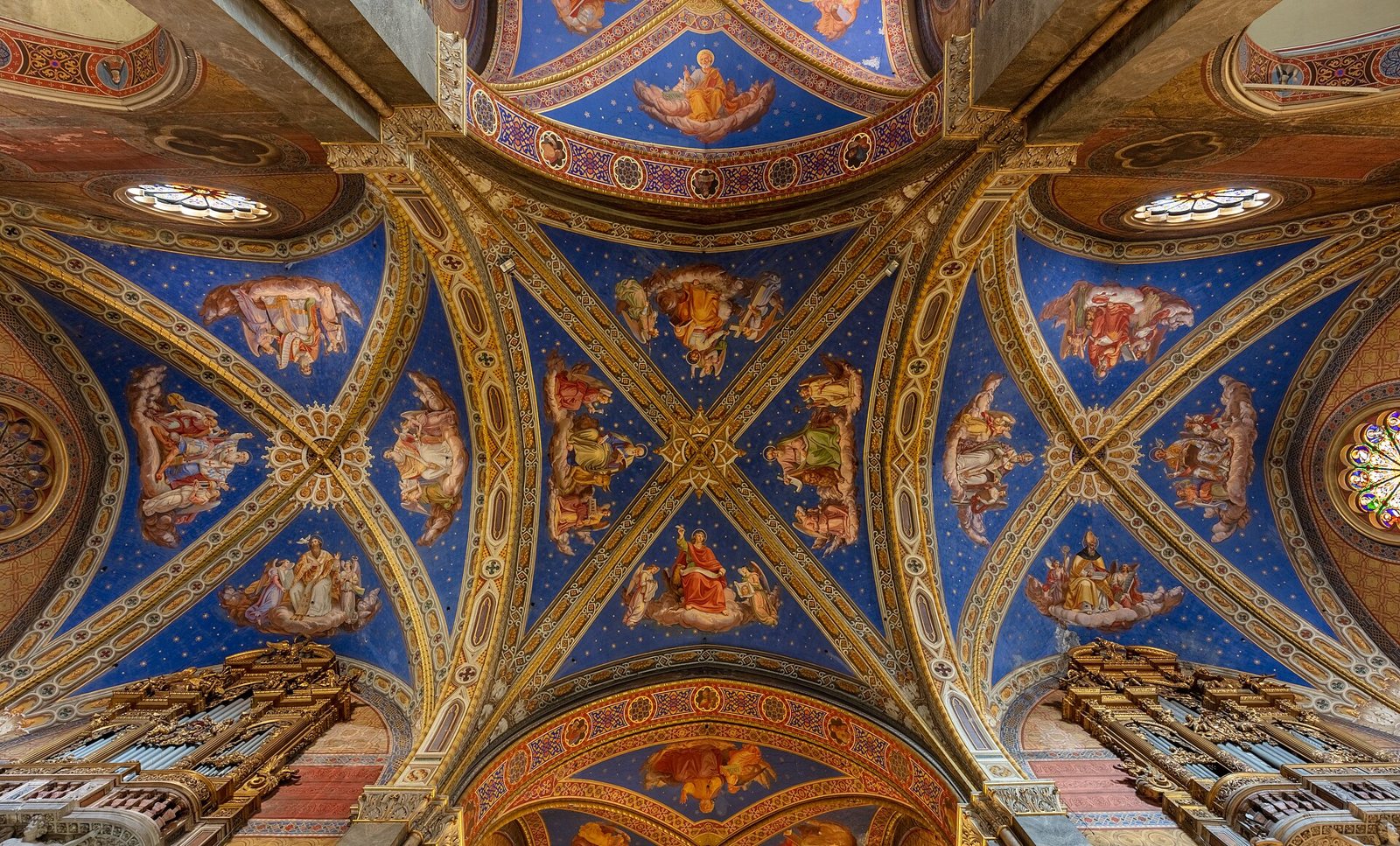 Looking up at a vaulted ceiling in Santa Maria sopra Minerva in Rome, painted deep blue with gold stars and gilded ribs, with painted figures in each panel and organ pipes at the lower corners