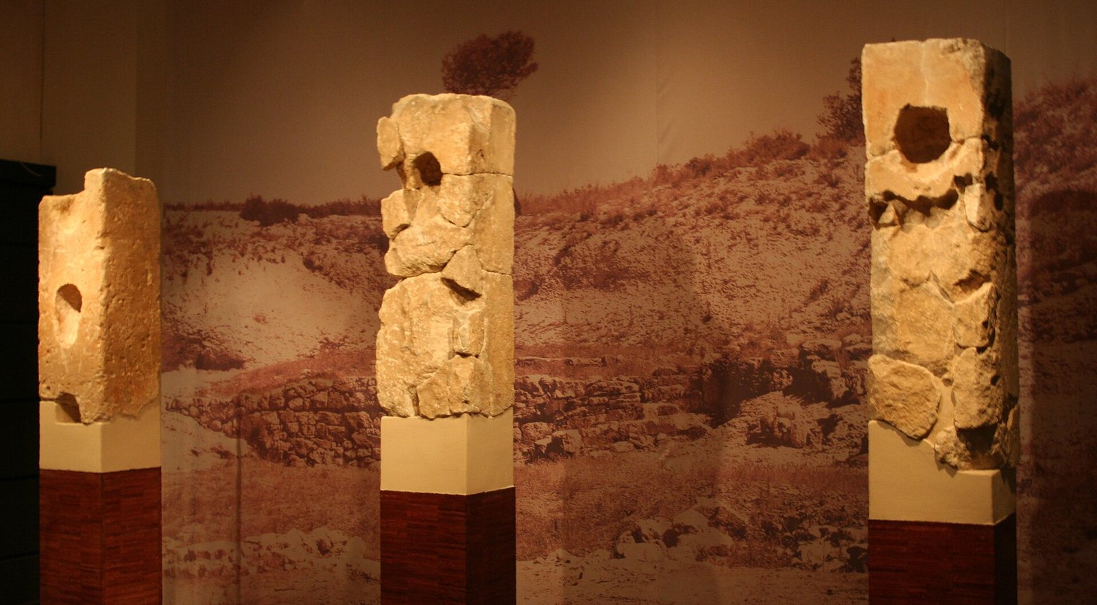 Three tall limestone pillars from the Roquepertuse sanctuary standing in a museum gallery, each with a round hollow cut into its face, in front of a large photographic backdrop of the hillside site