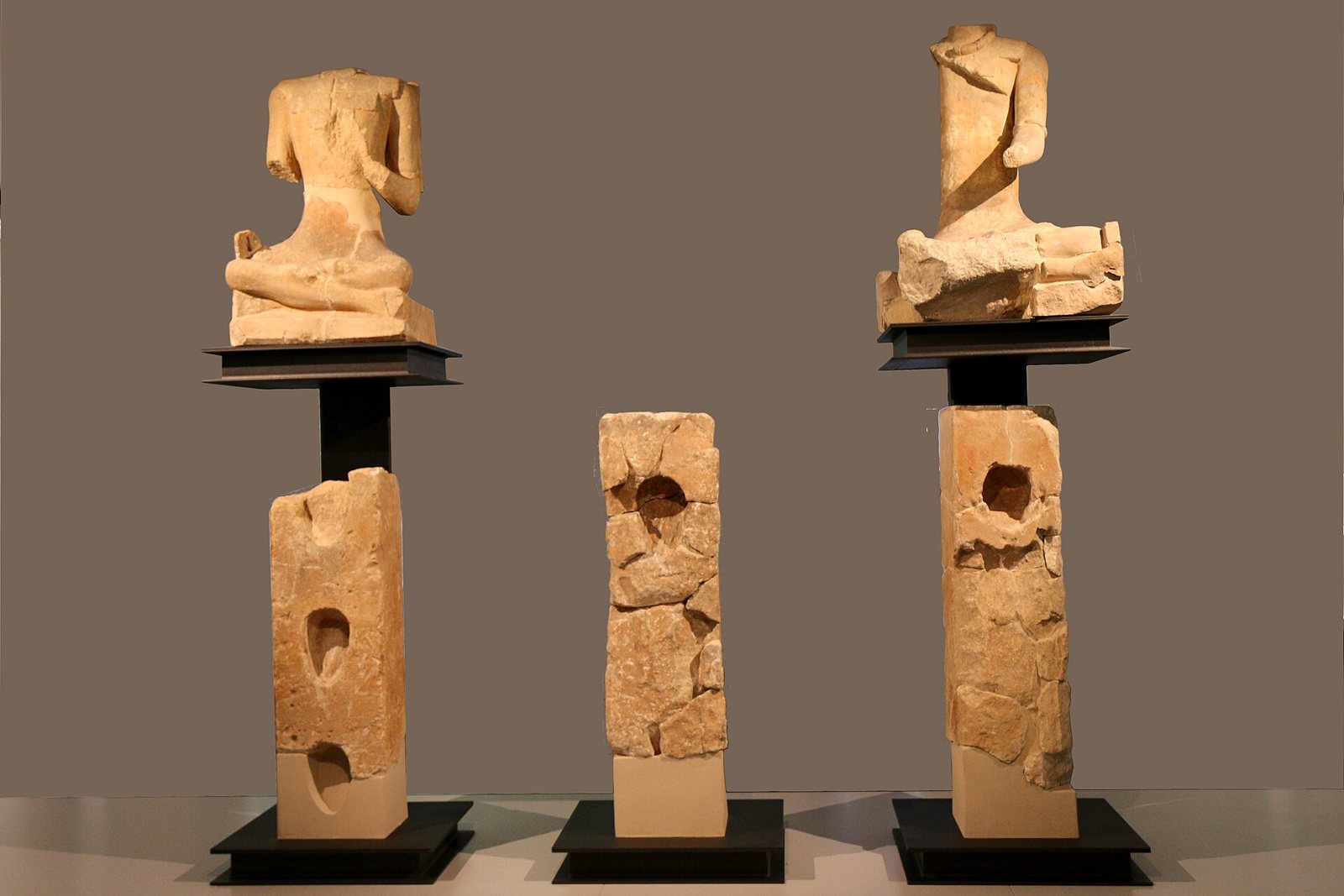 Two headless seated stone warriors on top of tall pillars with rounded hollows, and a third pillar between them, lit against a plain grey-brown backdrop in a museum