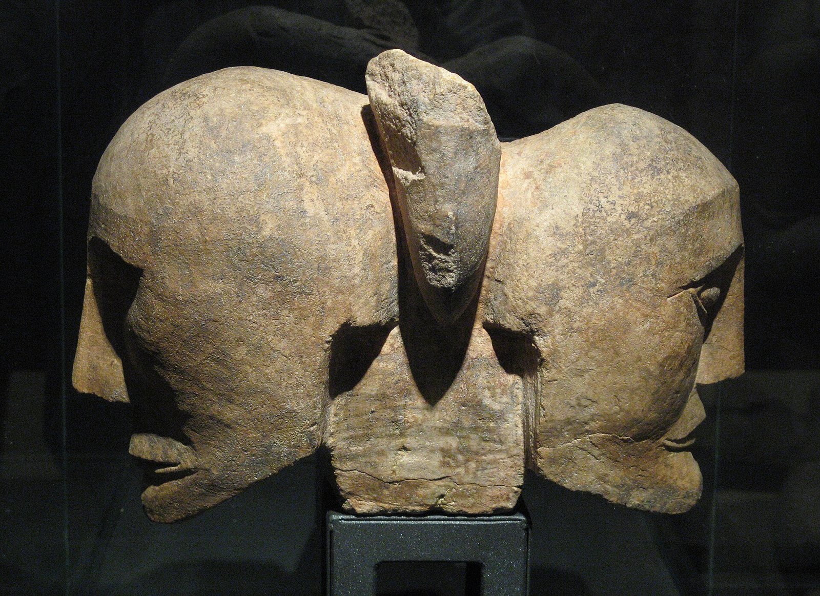 A pale stone sculpture of two heads joined back to back, one facing left and one right, with a narrow raised form rising between them, lit against a dark background