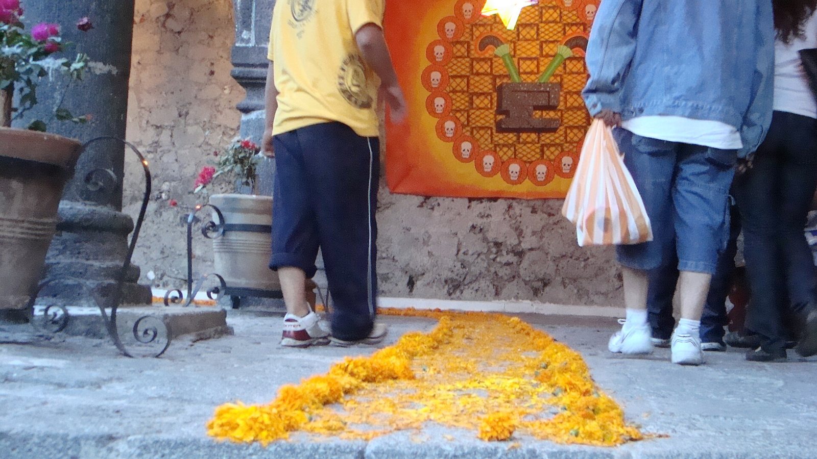 A trail of loose orange cempasúchil petals on a stone floor leading toward a wall hung with an orange banner bordered by small skulls, with visitors seen from behind