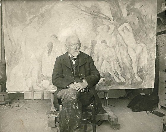Black-and-white photograph of an older white-bearded man in a dark suit seated in a studio, with a large unfinished canvas of bathing figures behind him