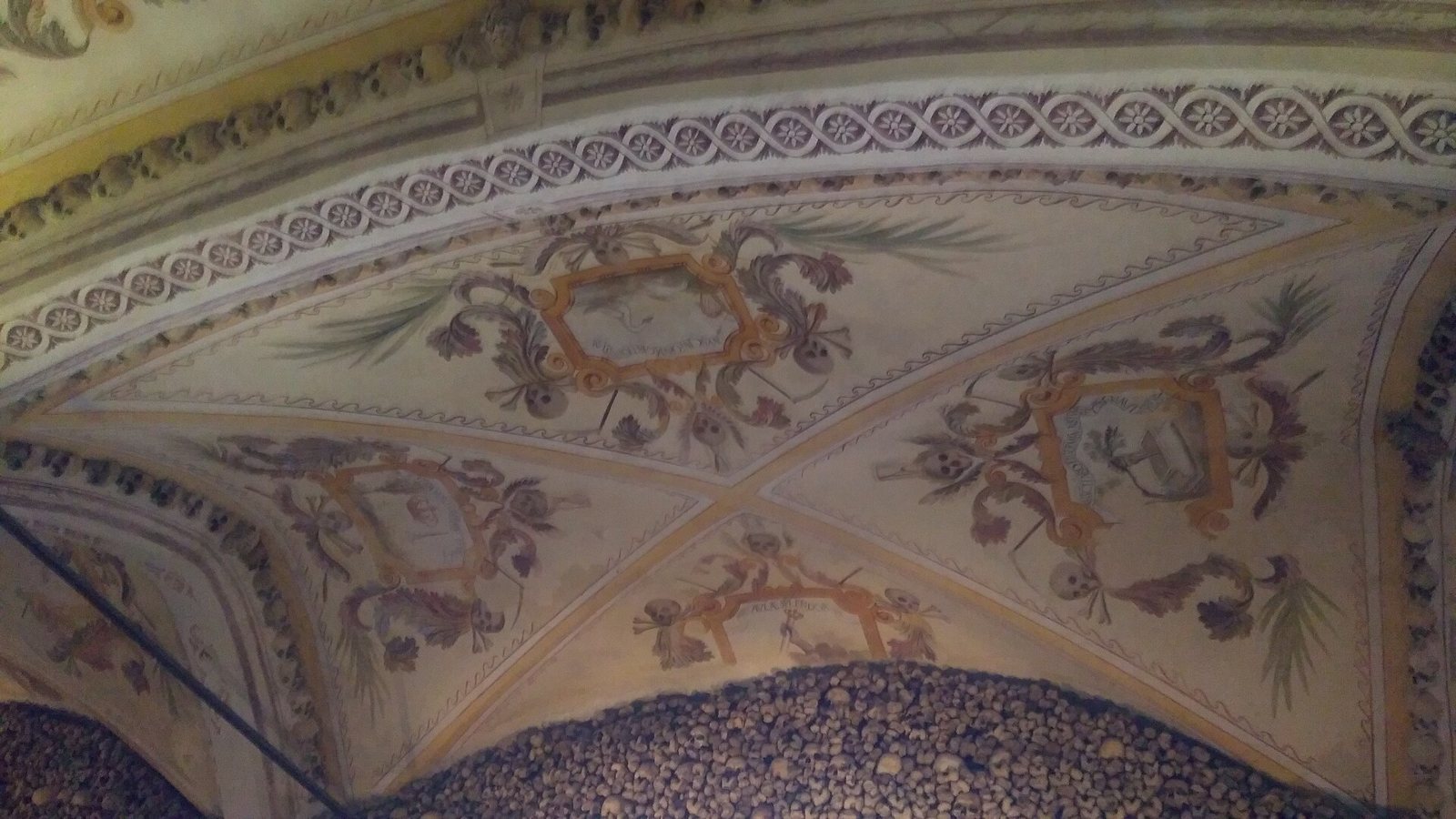 The vaulted ceiling of the Chapel of Bones painted with scrolling ornament and framed emblems, above walls lined with skulls