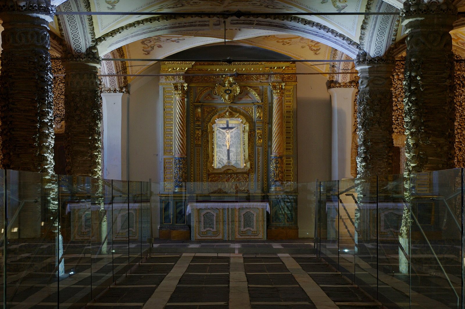 Inside the Capela dos Ossos in Évora: pillars and walls covered in skulls and bones frame a gilded altar with a crucifix beneath a painted vaulted ceiling, behind low glass screens