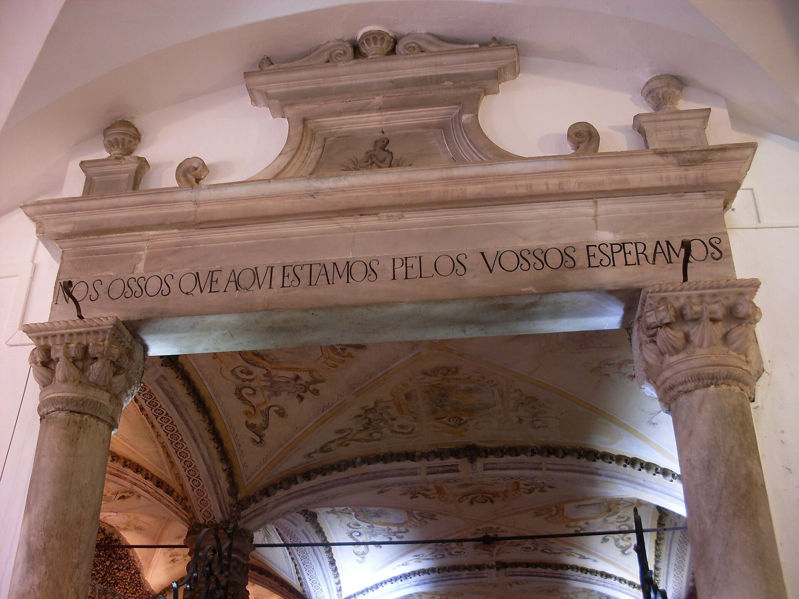 A carved stone lintel over the chapel entrance reading NOS OSSOS QUE AQUI ESTAMOS PELOS VOSSOS ESPERAMOS, with a painted vault beyond