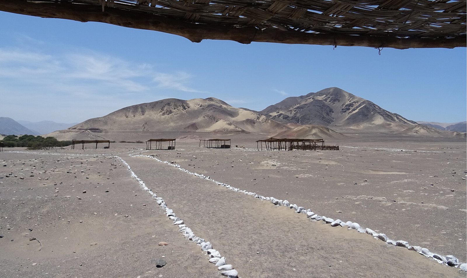 The Chauchilla cemetery in the Peruvian desert: a flat stony plain with lines of white stones marking paths, open-sided shelters in the distance and dry brown mountains beyond