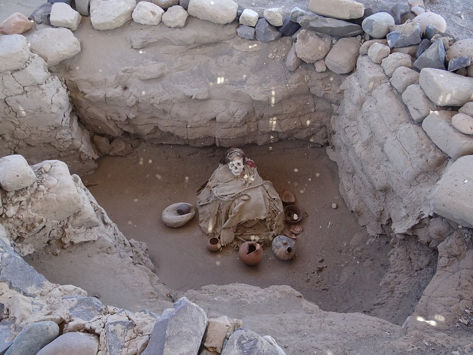 A single seated burial in a stone-edged pit at Chauchilla, wrapped in cloth, with small pottery bowls on the sand beside it