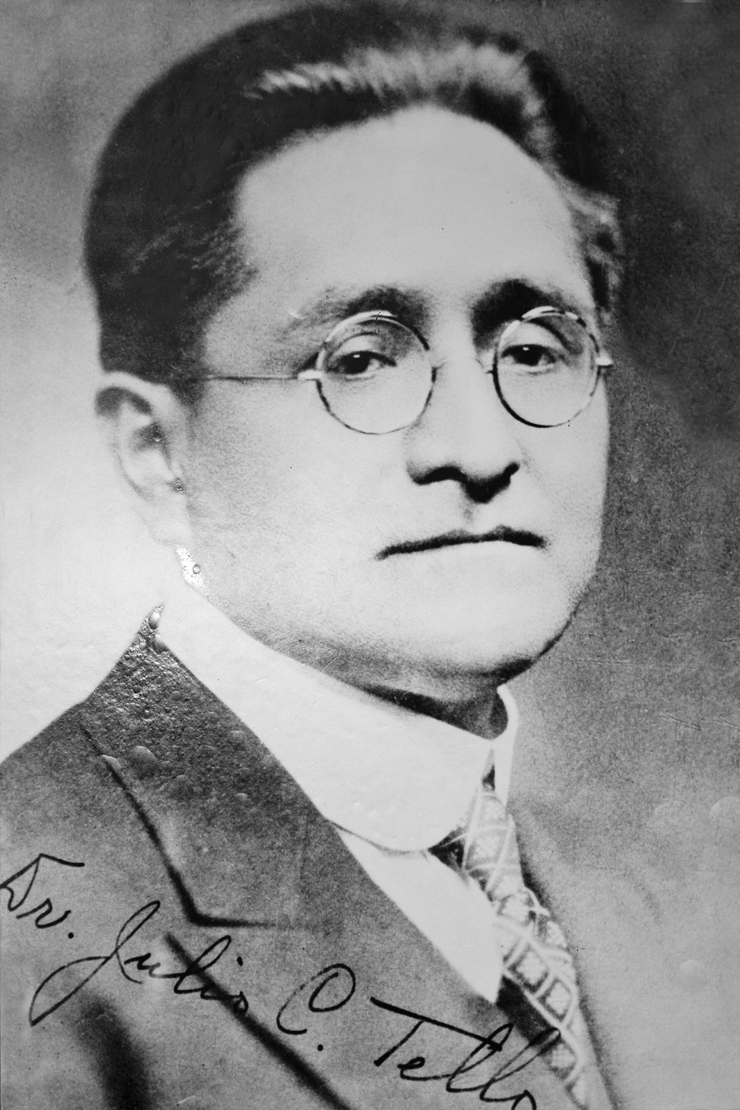 A black-and-white portrait of the archaeologist Julio C. Tello, a man in round glasses, a dark suit and a high white collar, with a signature along the bottom