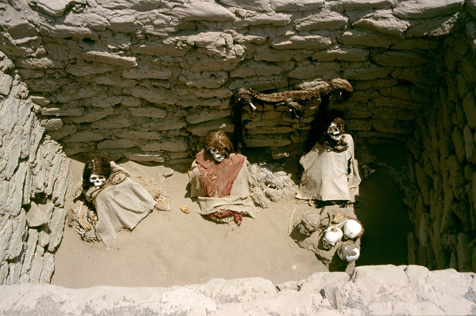 Three funerary bundles seated in an adobe-walled tomb at Chauchilla, wrapped in cloth and wearing red and cream textiles, with the tomb walls rising behind them
