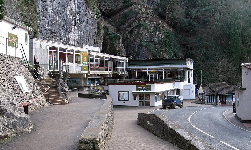 The entrance to Gough's Cave at the foot of a rock cliff in Cheddar Gorge, with a low white building, a road and a car in front