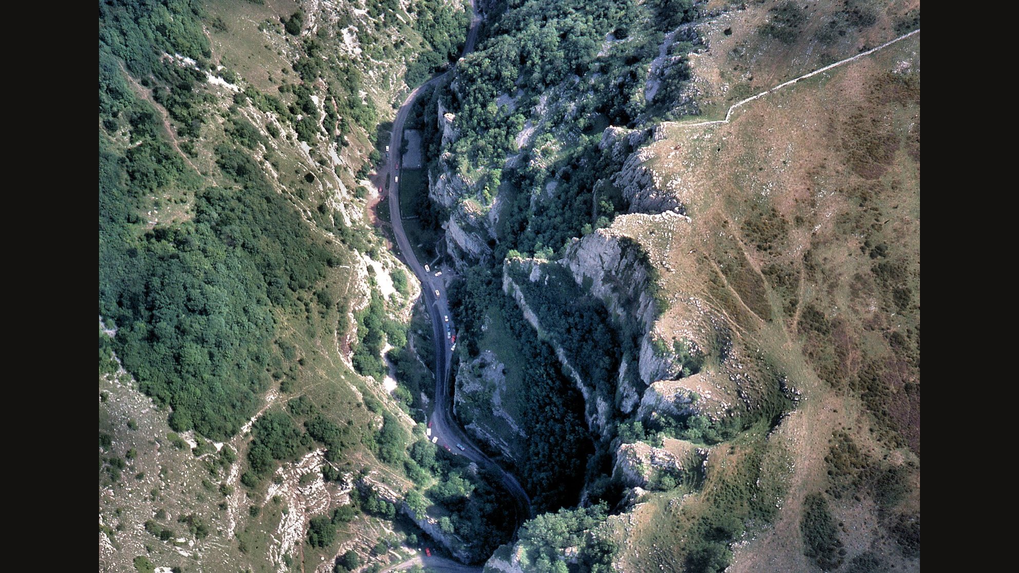 An aerial view of Cheddar Gorge in Somerset, a deep winding limestone gorge with a narrow road at the bottom and steep green and grey slopes on both sides
