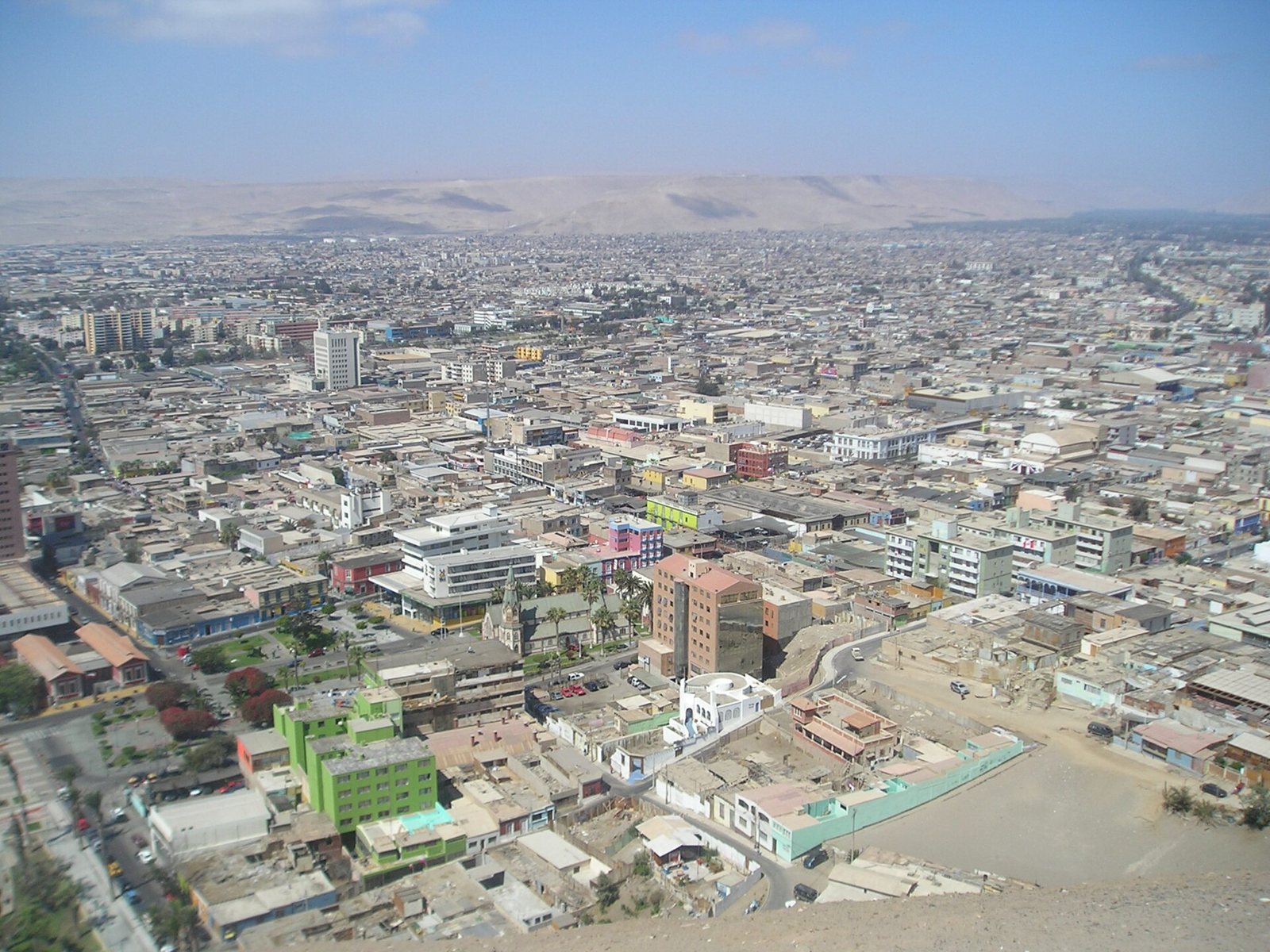 A view east over the city of Arica from El Morro: dense low buildings stretching toward brown desert hills, with a church and green and pink buildings in the middle distance