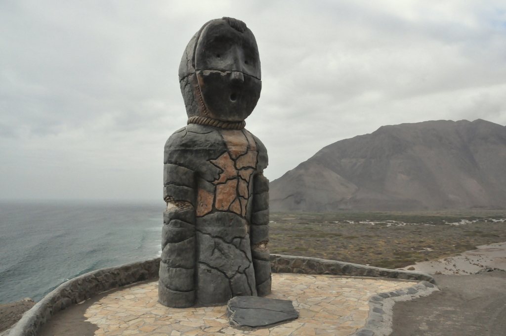 A weathered sculpture of a standing figure with a mask-like face and a rope collar on a paved lookout above the sea, with a brown desert mountain and a grey sky behind