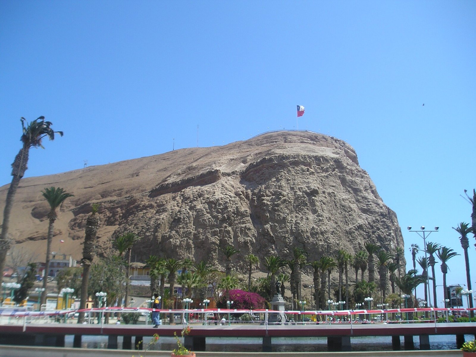 The Morro de Arica, a tall brown rock headland with a Chilean flag on top, seen over palm trees and a promenade under a clear blue sky
