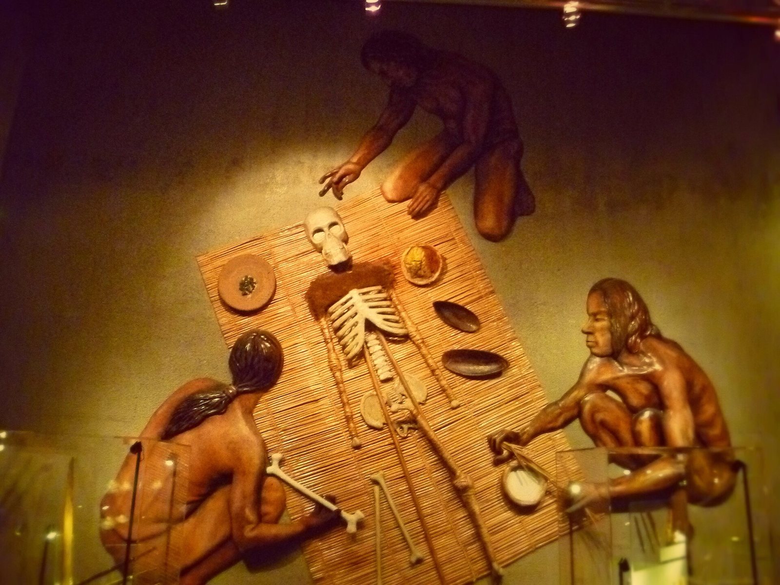 A museum wall relief of three figures kneeling around a skeleton laid on a reed mat, one holding a bone, with bowls of pigment and ash beside it