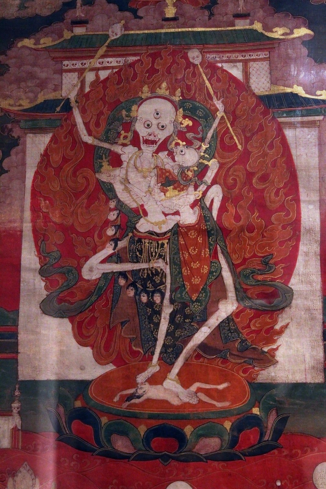 A detail of a Tibetan painting on cloth showing a pair of white skeleton figures with three eyes and crowns, holding staffs and dancing on a lotus seat against a red halo of flames