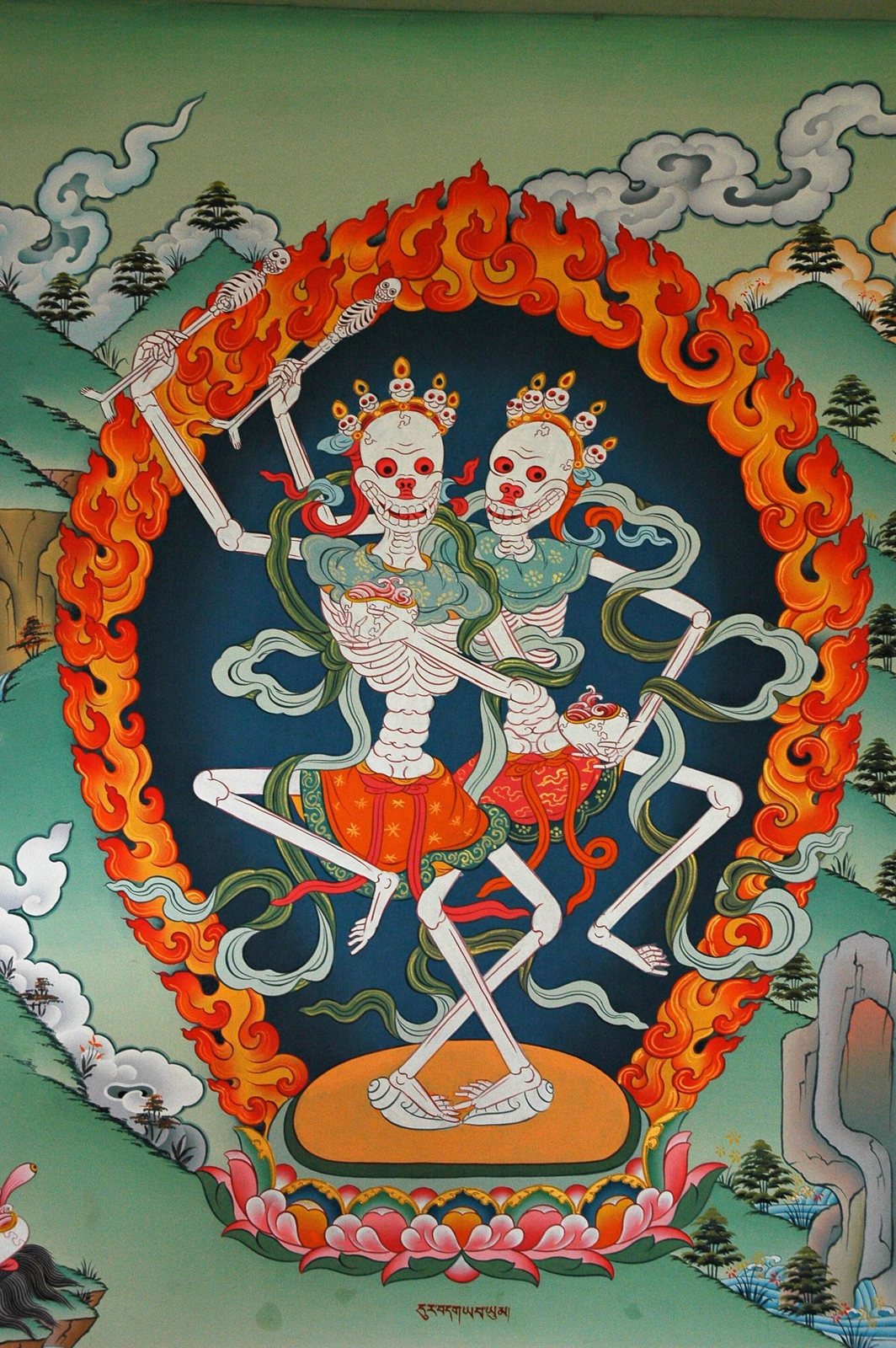 A wall painting of two white skeletons with skull crowns dancing together with legs interlocked, inside a ring of orange flames, on a lotus base among green hills and clouds