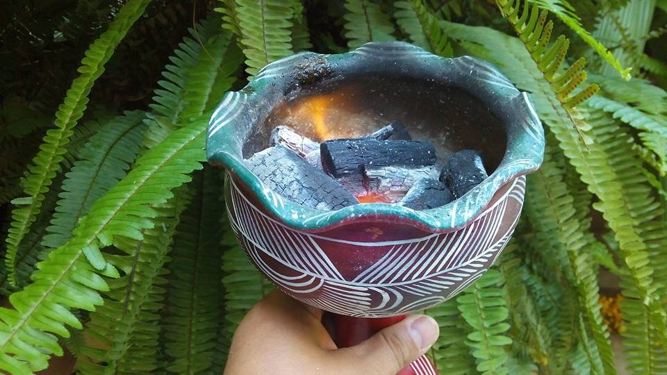 Una mano sostiene un incensario de barro vidriado en verde y decorado con líneas blancas incisas, con carbón encendido adentro y hojas de helecho al fondo