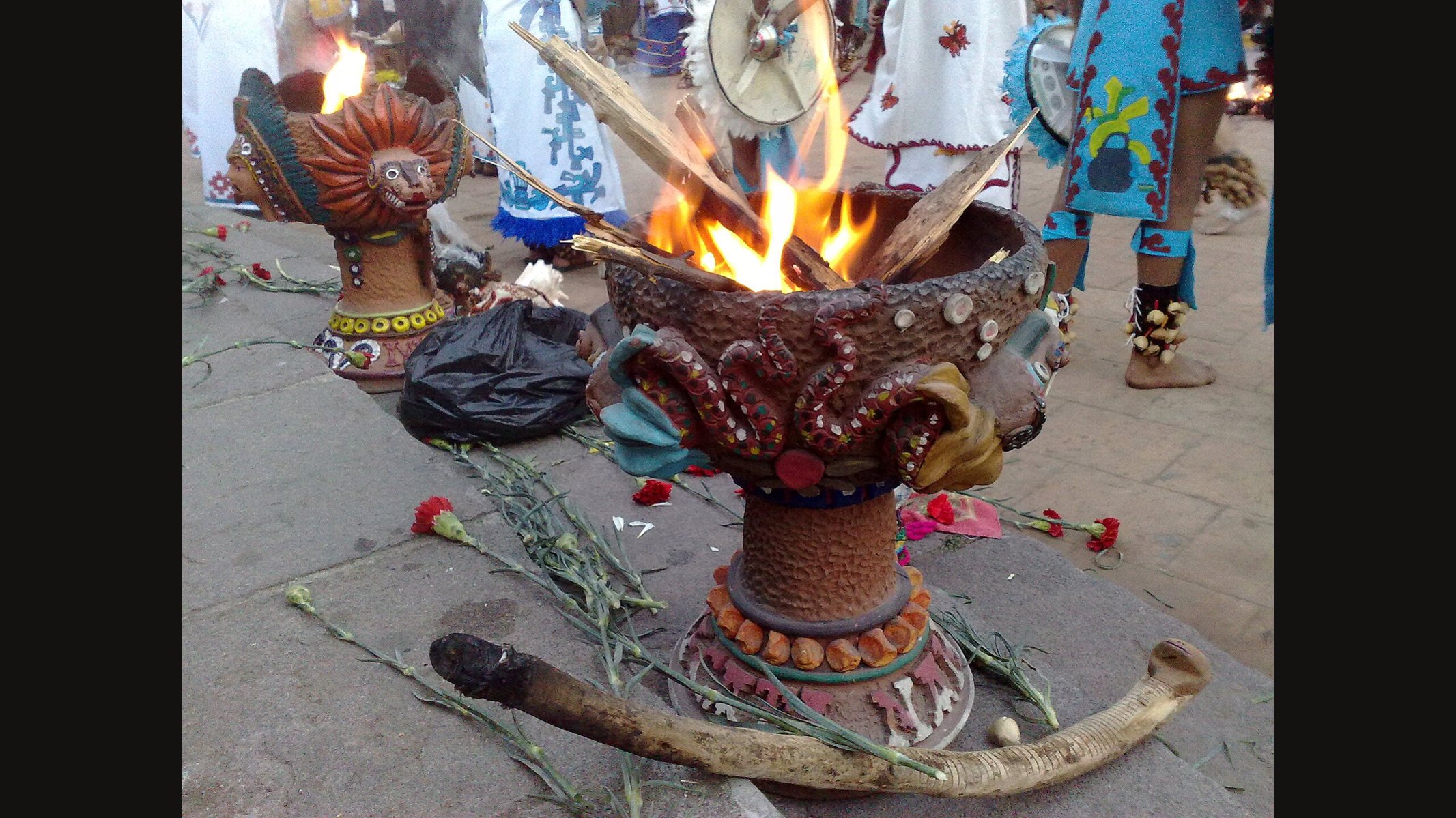 Un gran sahumerio de barro decorado con llamas que salen de la leña en su interior, claveles rojos sobre la piedra y otro sahumerio con forma de cabeza con plumas a un lado