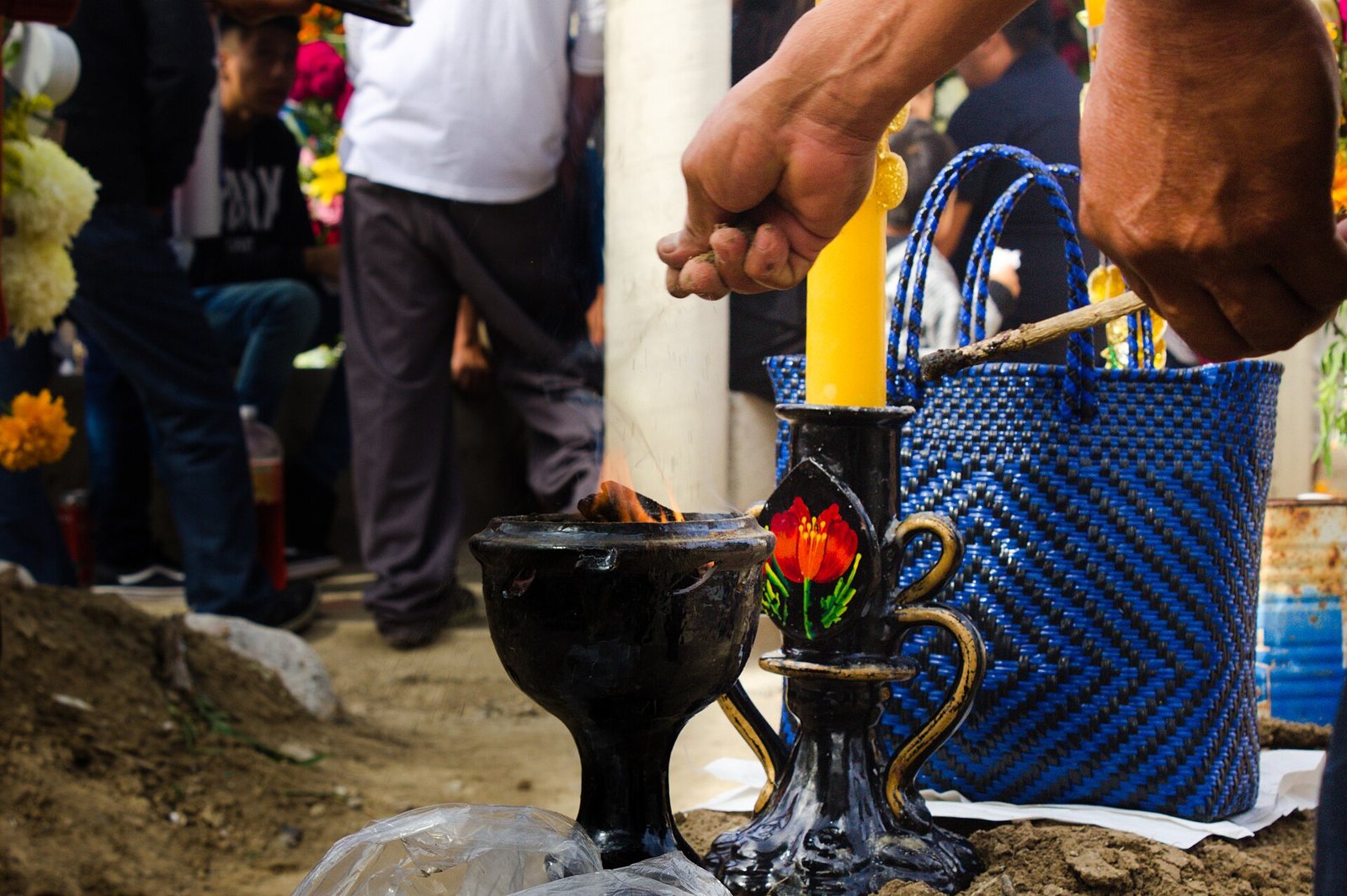 Las manos de un hombre mueven el copal en un incensario de barro negro junto a una vela amarilla encendida en una tumba de Día de Muertos, con una bolsa tejida azul y gente de pie detrás
