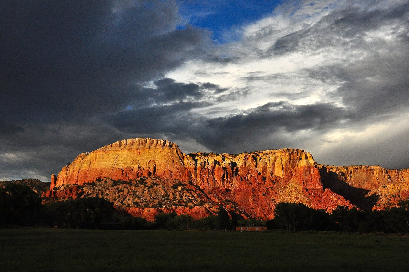 Red and yellow sandstone cliffs lit by low sun beneath dark storm clouds, above a green field