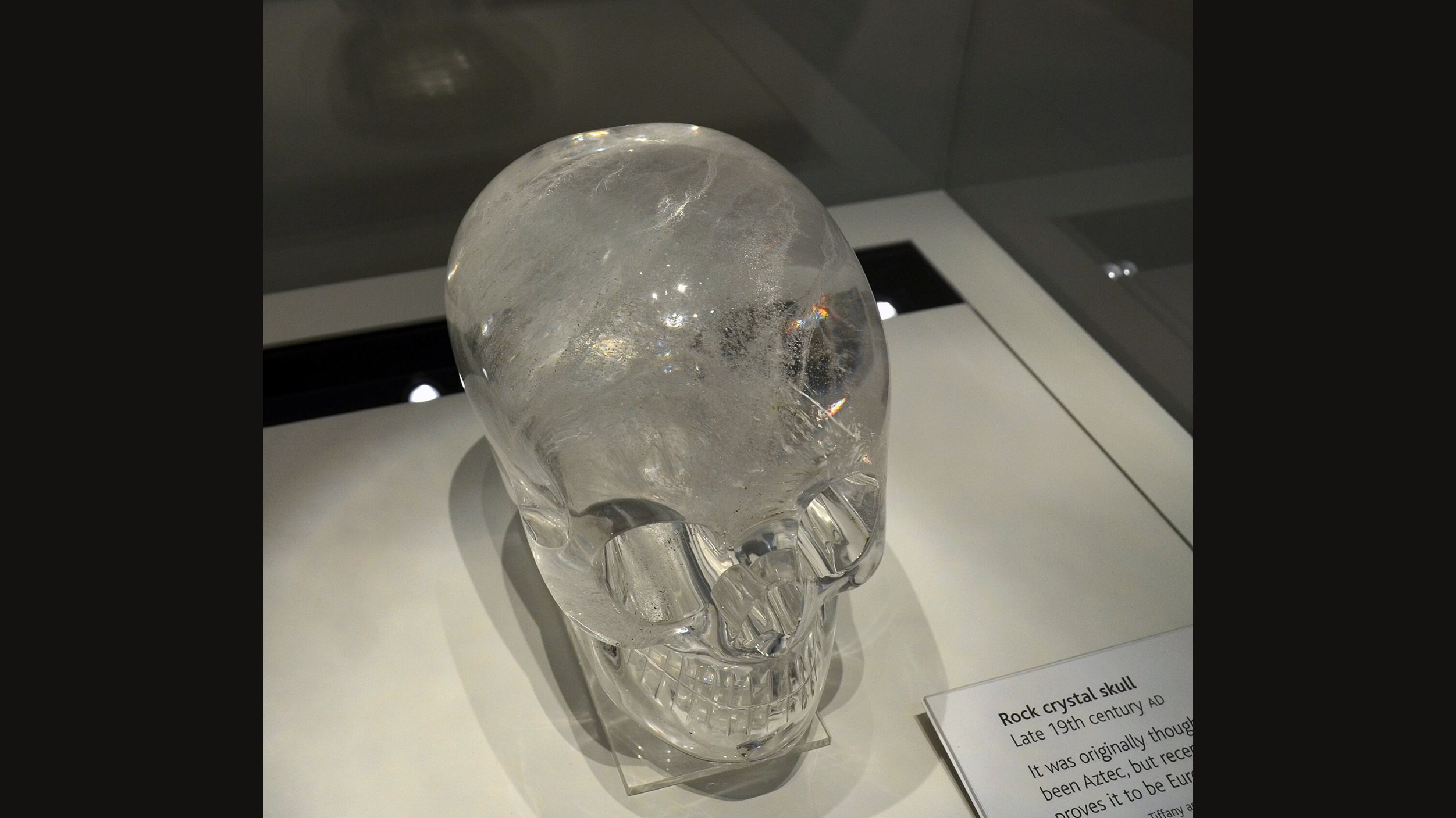 The British Museum's rock crystal skull seen from above and the front, clear quartz on a pale display plinth, with part of its museum label visible