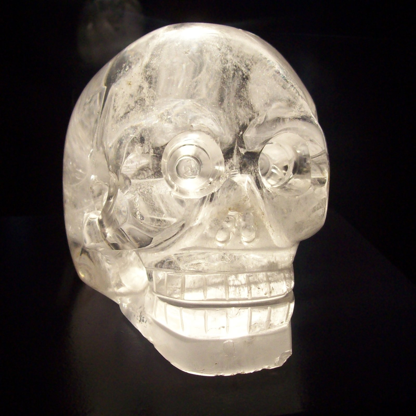 A small clear quartz skull photographed against black, with round carved eyes and a row of carved teeth