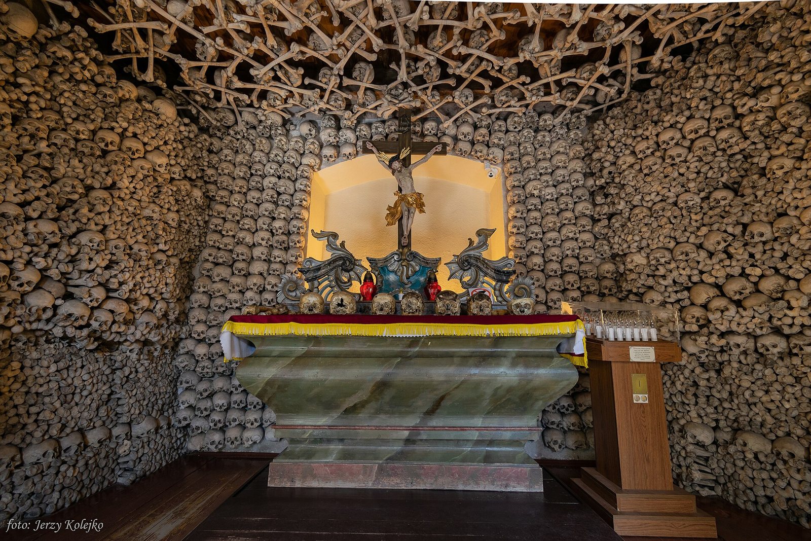 The chapel altar, a green marbled table with a red and gold altar cloth, beneath a lit niche holding a crucifix, all set against walls and a ceiling covered in skulls and bones
