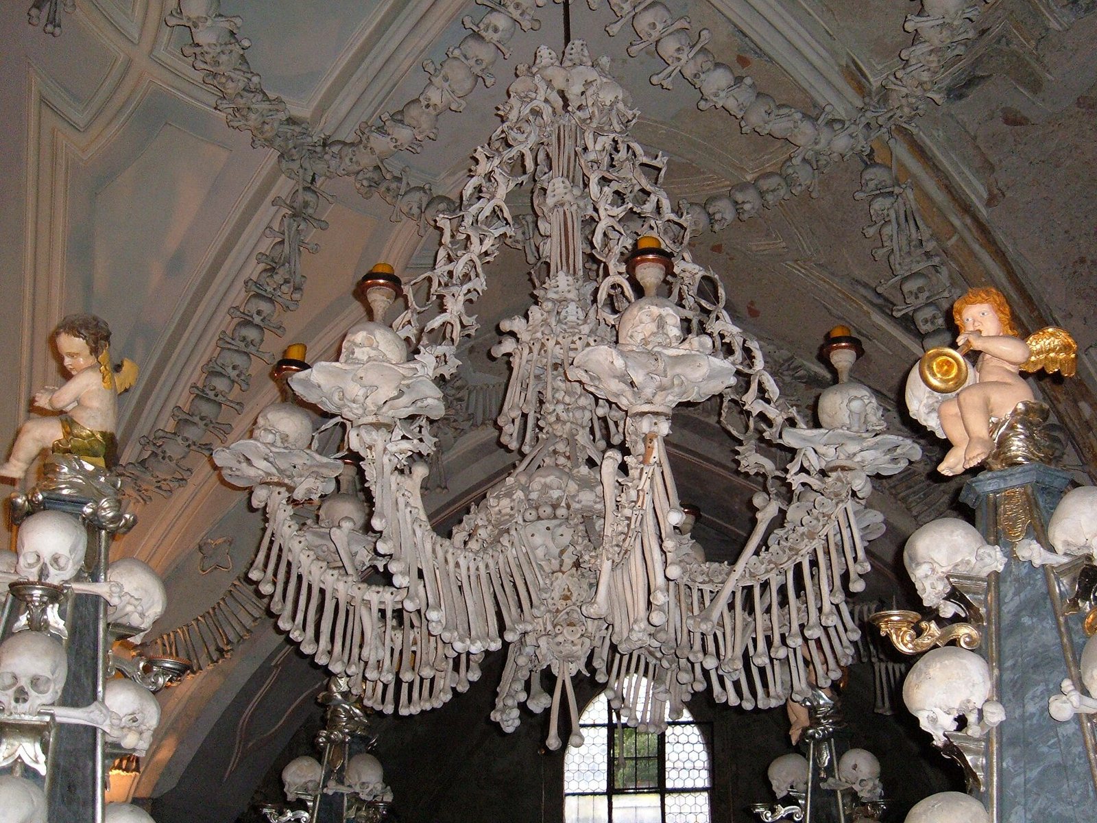 A chandelier made of human bones hanging in the Sedlec Ossuary, with skull-topped pillars and gilded cherubs on either side and garlands of skulls along the vaulted ceiling