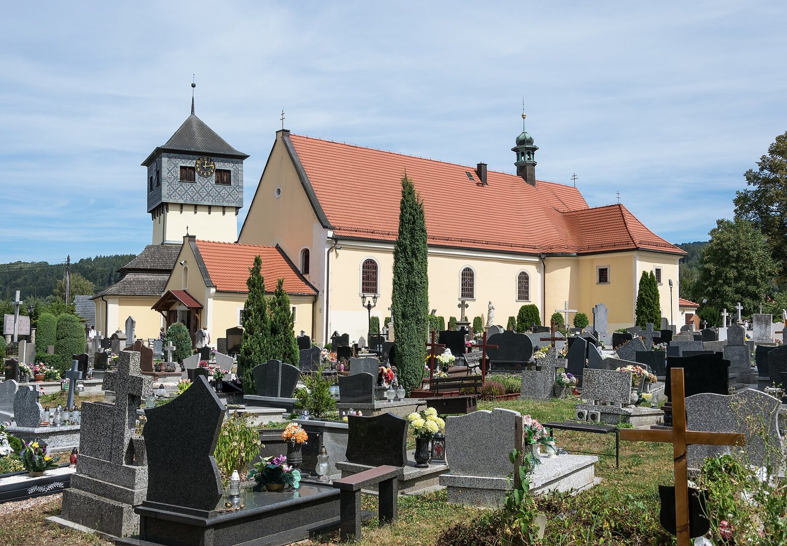 A cream-walled church with a red tiled roof and a slate-clad bell tower, seen across a cemetery of dark granite headstones, crosses and flowers