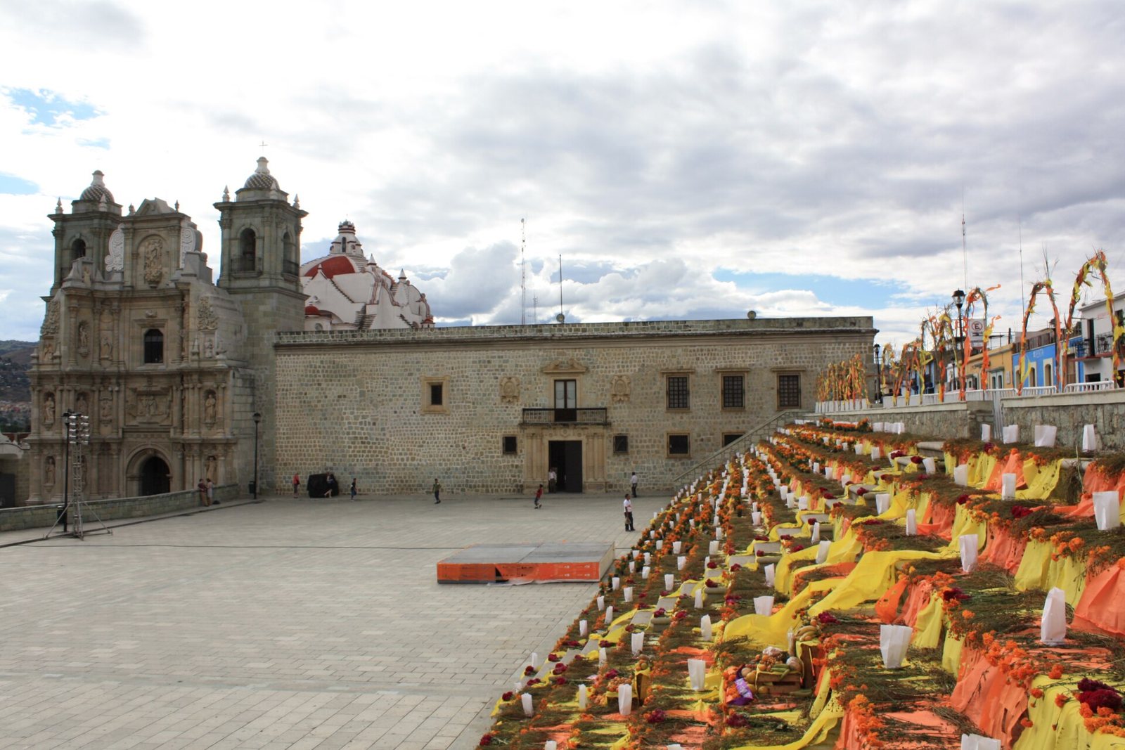 A long row of Día de Muertos altars with candles, marigolds and orange and yellow cloth laid along the steps of a plaza in Oaxaca de Juárez, with a carved stone church and colonial buildings behind