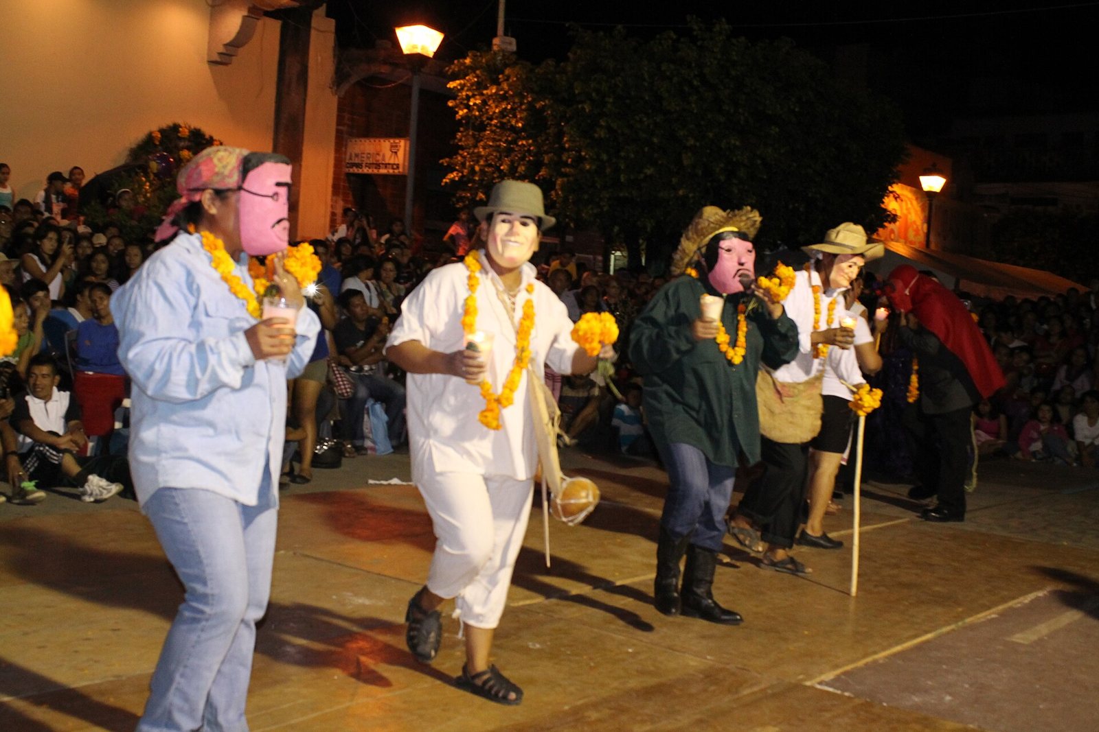 Masked Xantolo dancers in white and green clothes, straw hats and marigold garlands, carrying canes across a lit street at night while a crowd watches