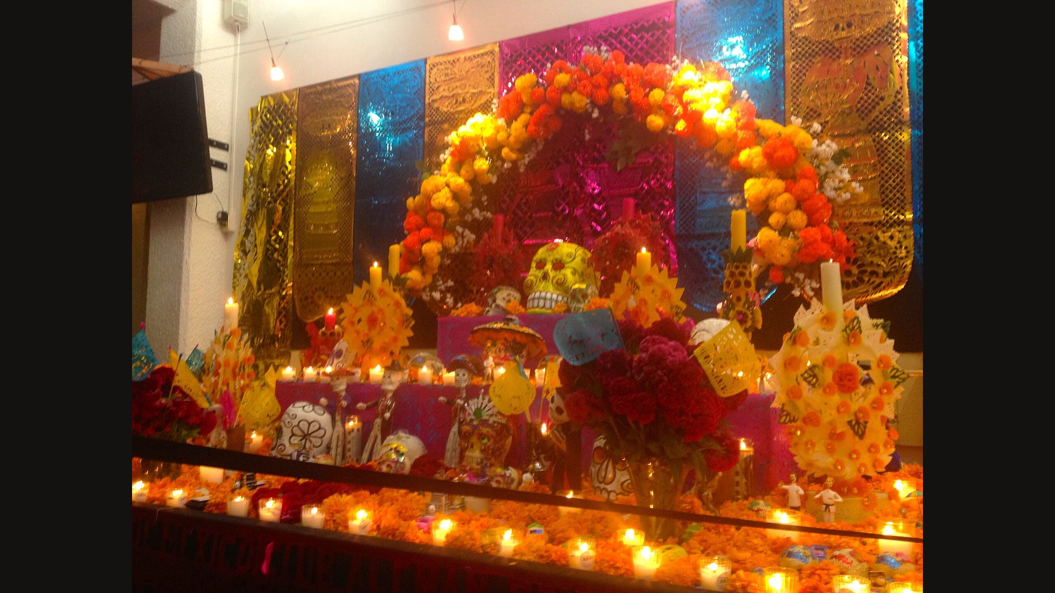 A tiered Día de Muertos ofrenda in Mexico City: an arch of orange and yellow marigolds, a painted sugar skull at the top, rows of lit candles, red cockscomb flowers and bright papel picado hanging behind