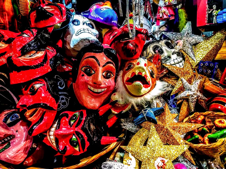 A market display crowded with painted papier-mâché masks of red devils, smiling faces and white skulls, beside glittering gold and silver paper stars