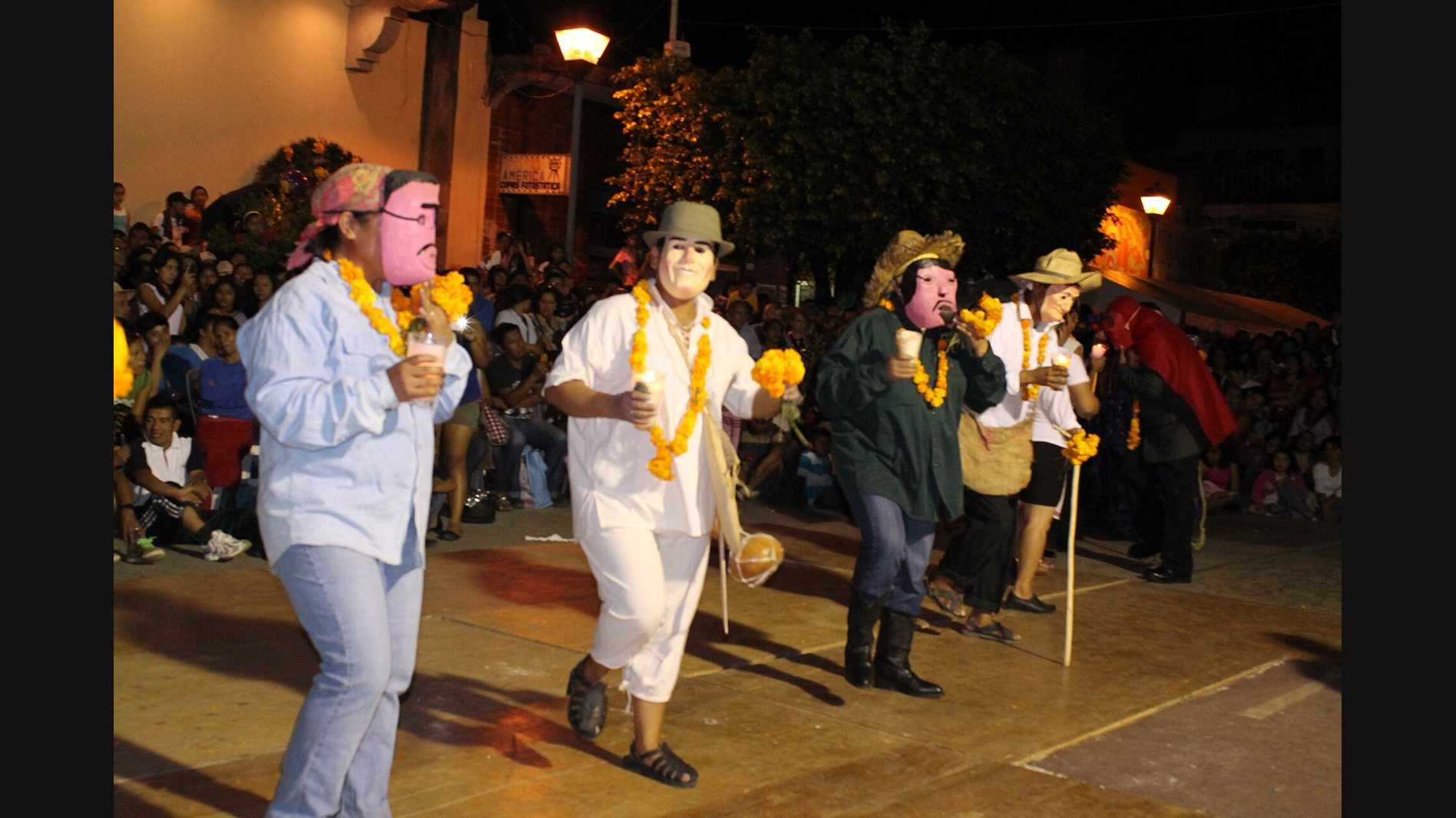Xantolo dancers at night in pale and pink masks, hats and marigold garlands, moving through a square in front of a seated crowd