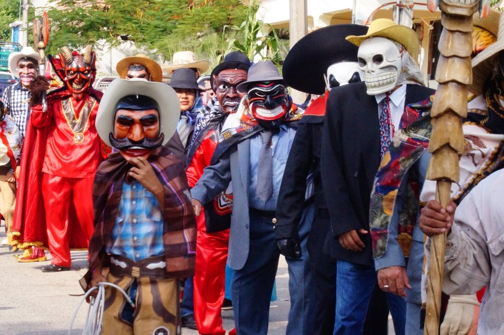 A line of masked Xantolo characters in a street: a cowboy, red devils with horns, dark-faced masks under wide hats and a white skull mask, with a carved staff at the right