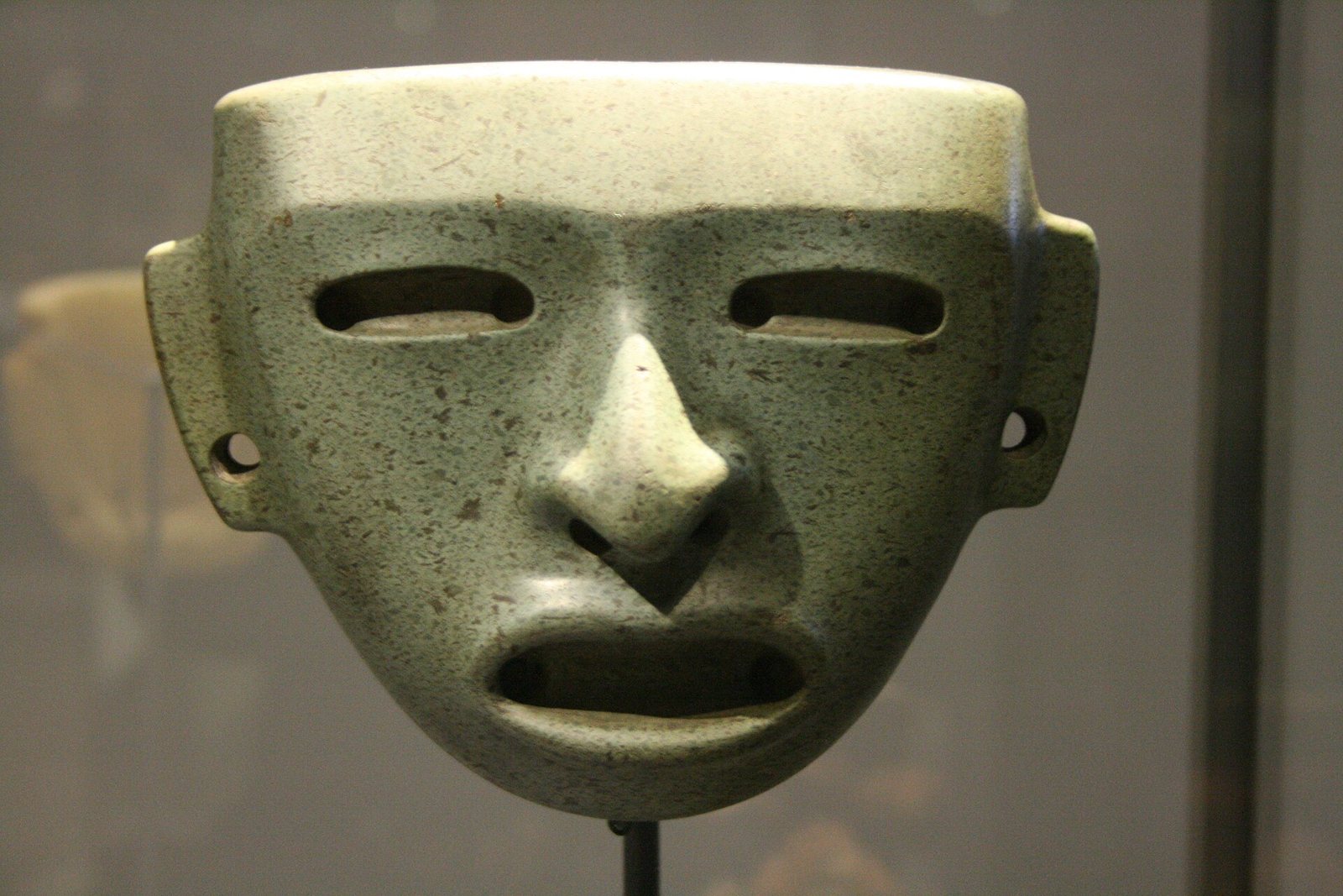 A smooth green speckled stone mask with slit eyes, a broad nose, an open mouth and pierced ear tabs, mounted on a stand in a museum case