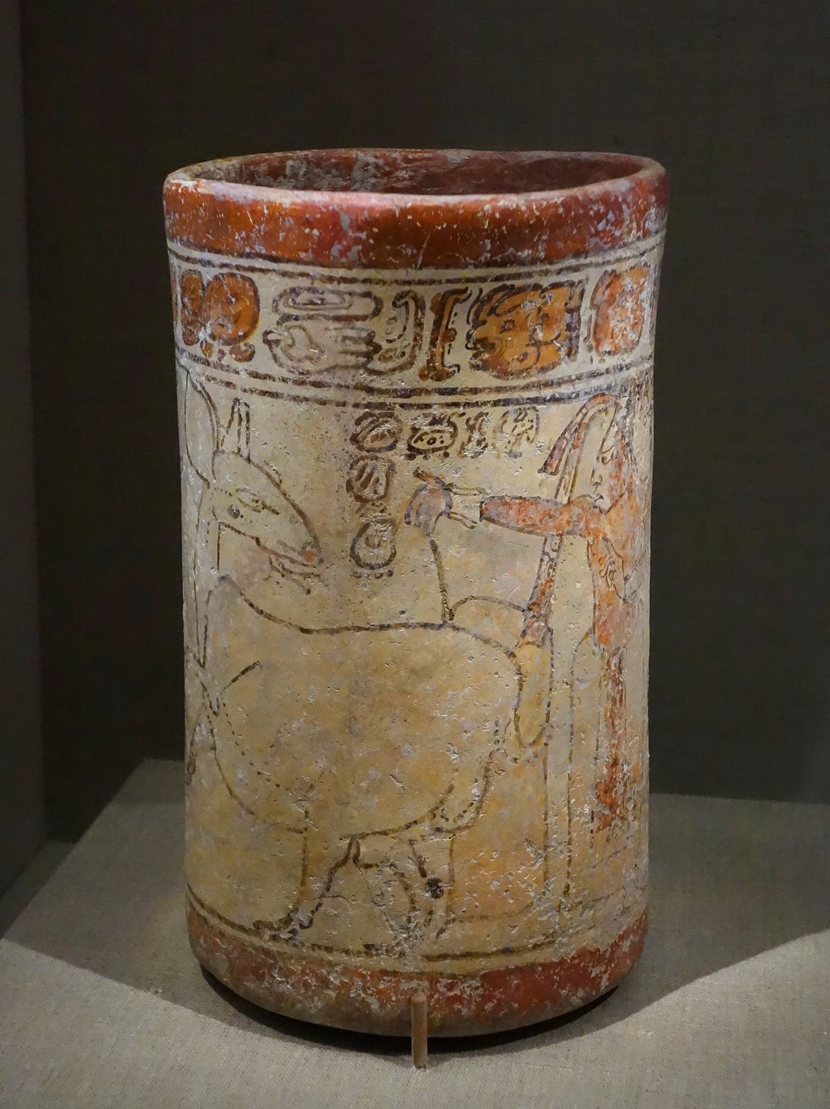 A tall cylinder vase with a band of hieroglyphs at the top and a drawn figure beside a deer, in red and cream