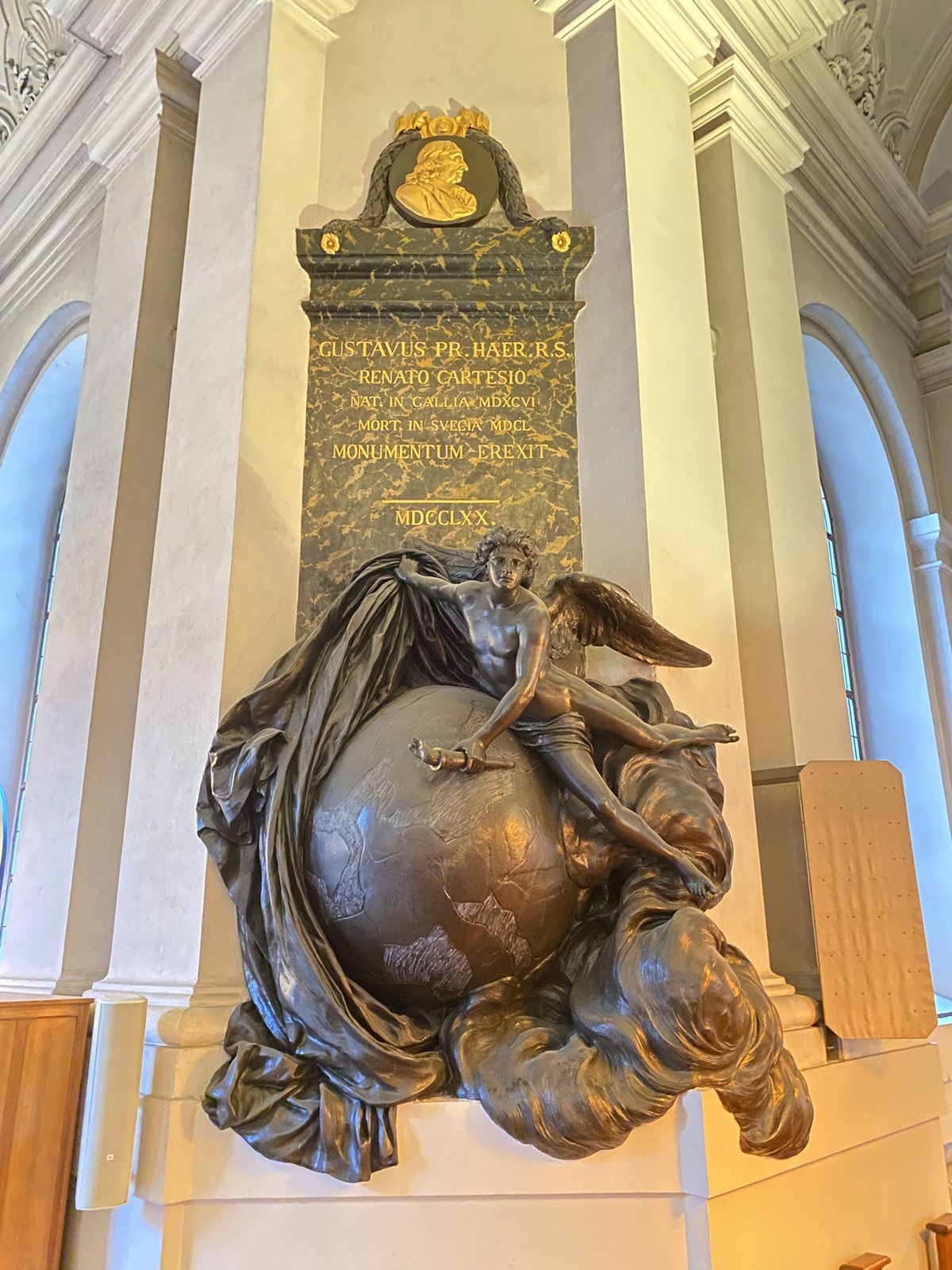 A dark bronze winged figure sprawled over a globe below a tall black marble panel with a gold relief portrait and a Latin inscription, in a white church interior