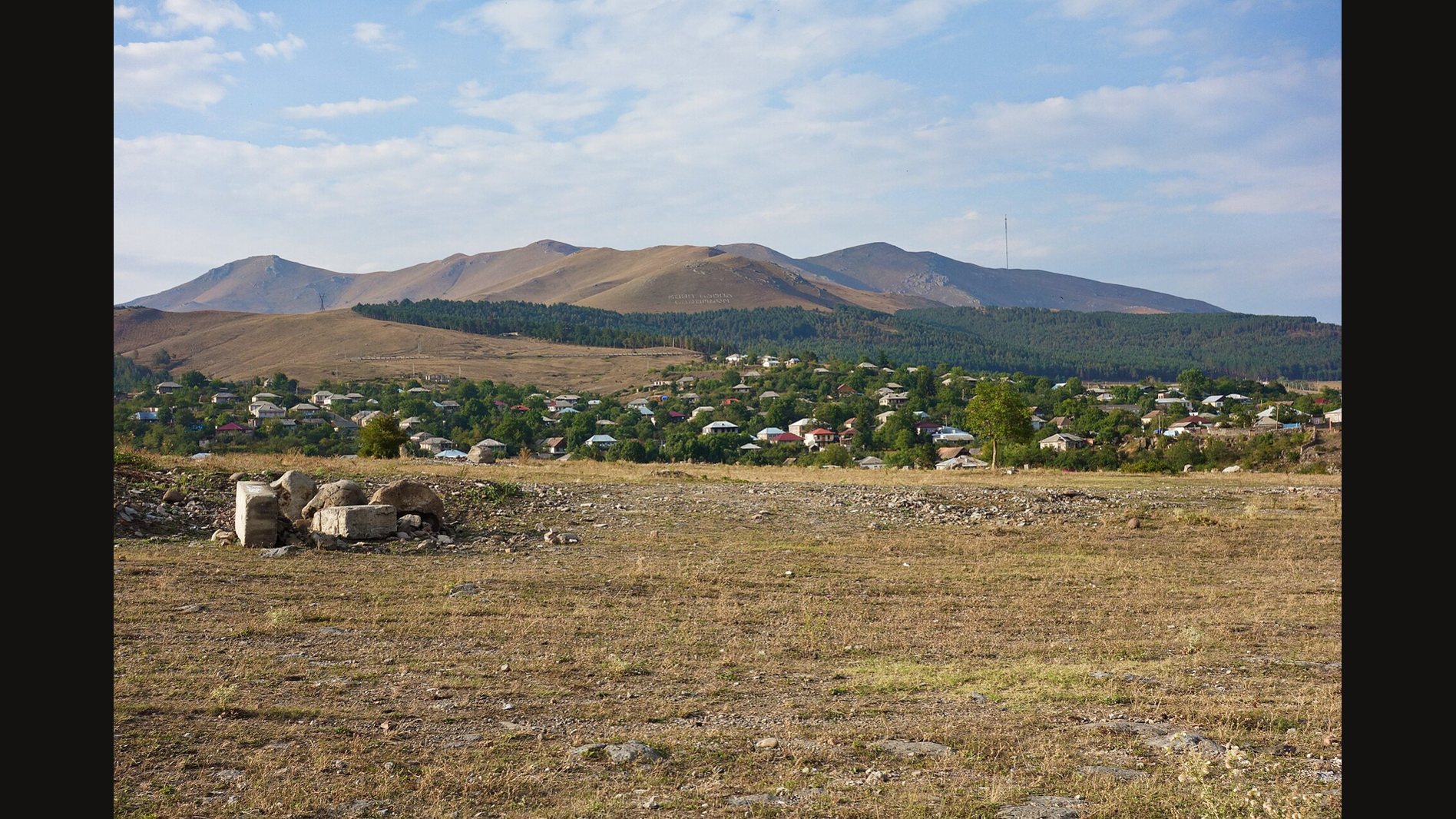 The town of Dmanisi among green trees below a long mountain ridge, with dry open grassland and a few pale stone blocks in the foreground