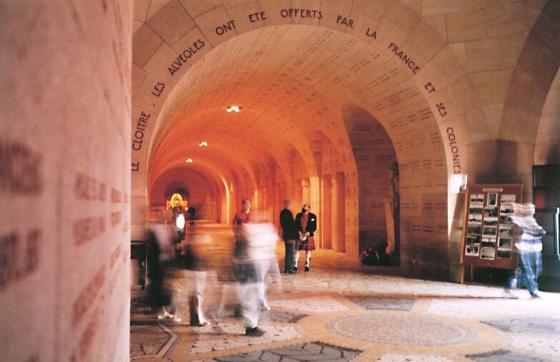 A long vaulted stone cloister glowing orange, with names carved into the arches and walls, and a few visitors blurred by movement along the floor