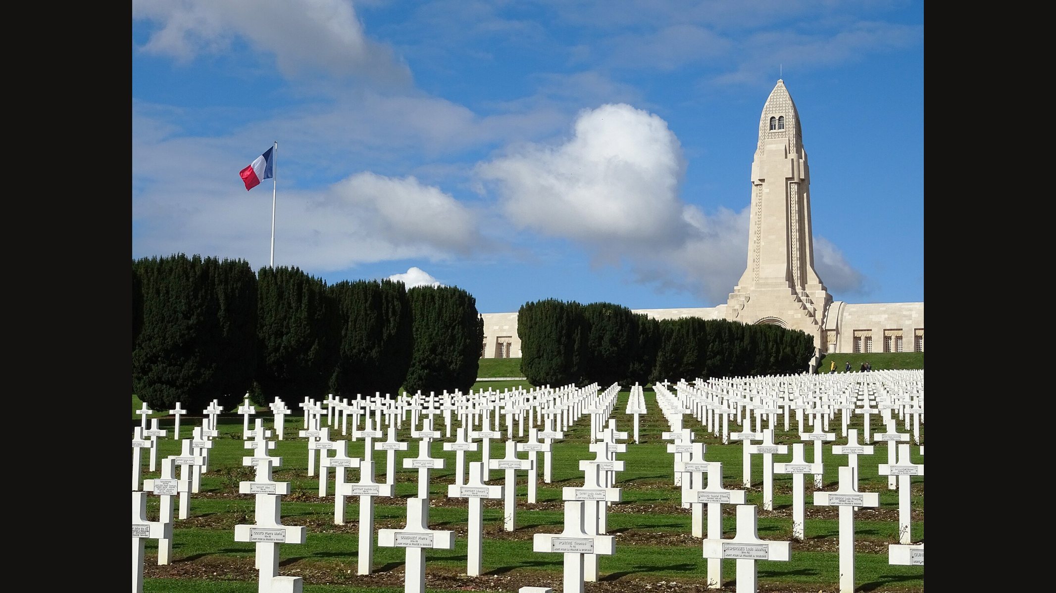 The Douaumont ossuary under a blue sky with a French flag: a tall pale stone lantern tower above a long low building, with rows of white crosses in the cemetery in front
