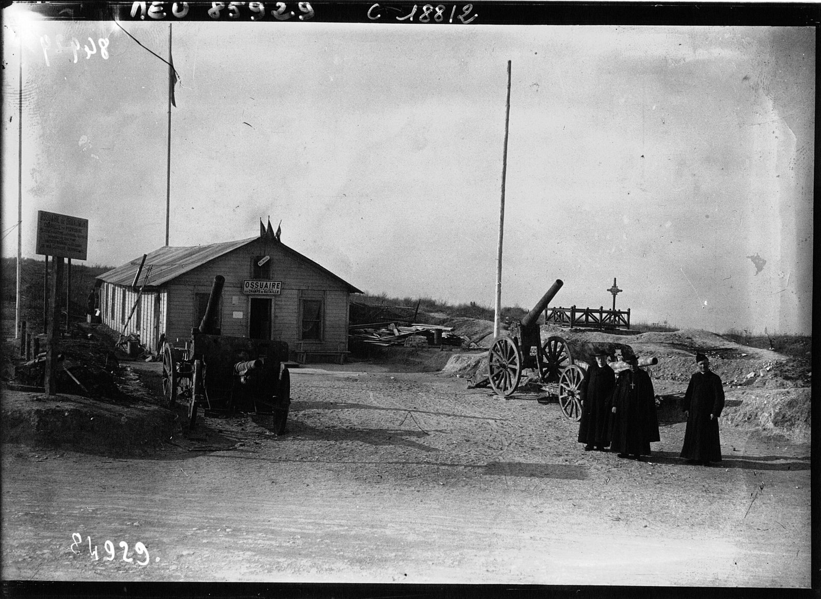 A black-and-white photograph of the temporary ossuary at Douaumont: a wooden hut with a sign, a field gun and a cross on bare ground, with three clergy in dark cassocks standing at the right