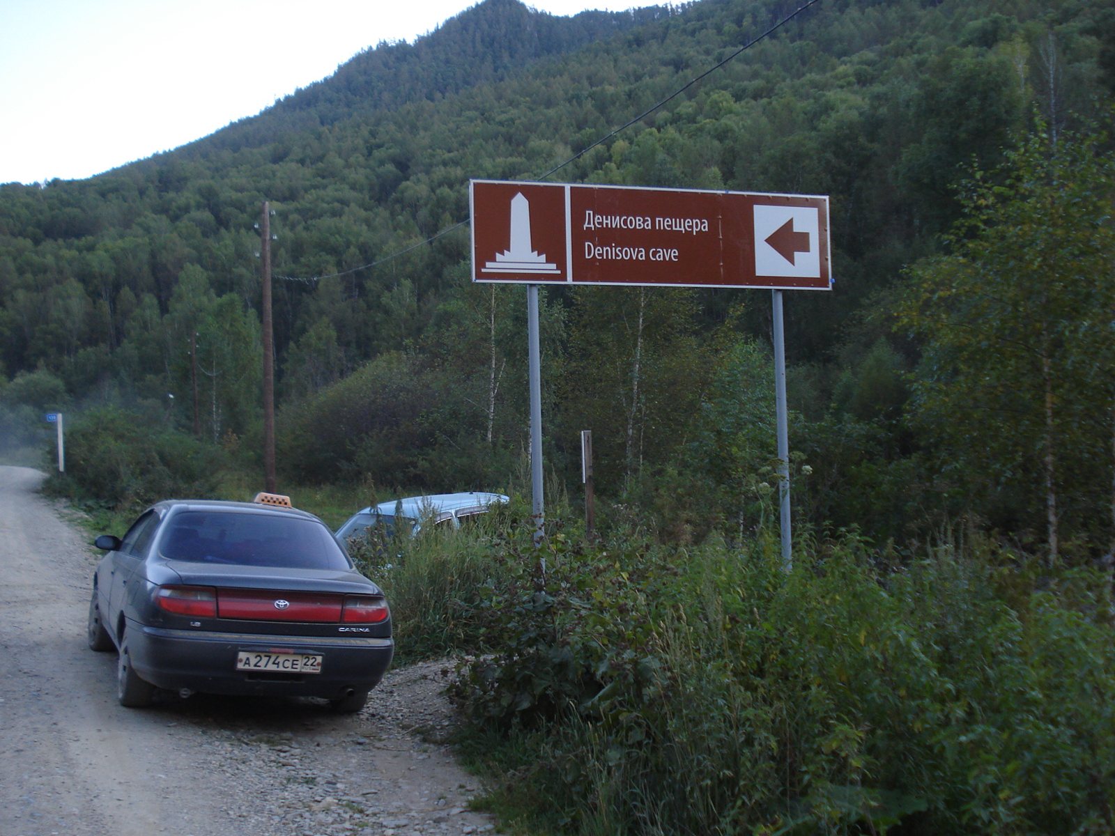Gravel track through forested hills in the Altai Mountains with a brown road sign reading Denisova cave in Russian and English and a car parked at the roadside