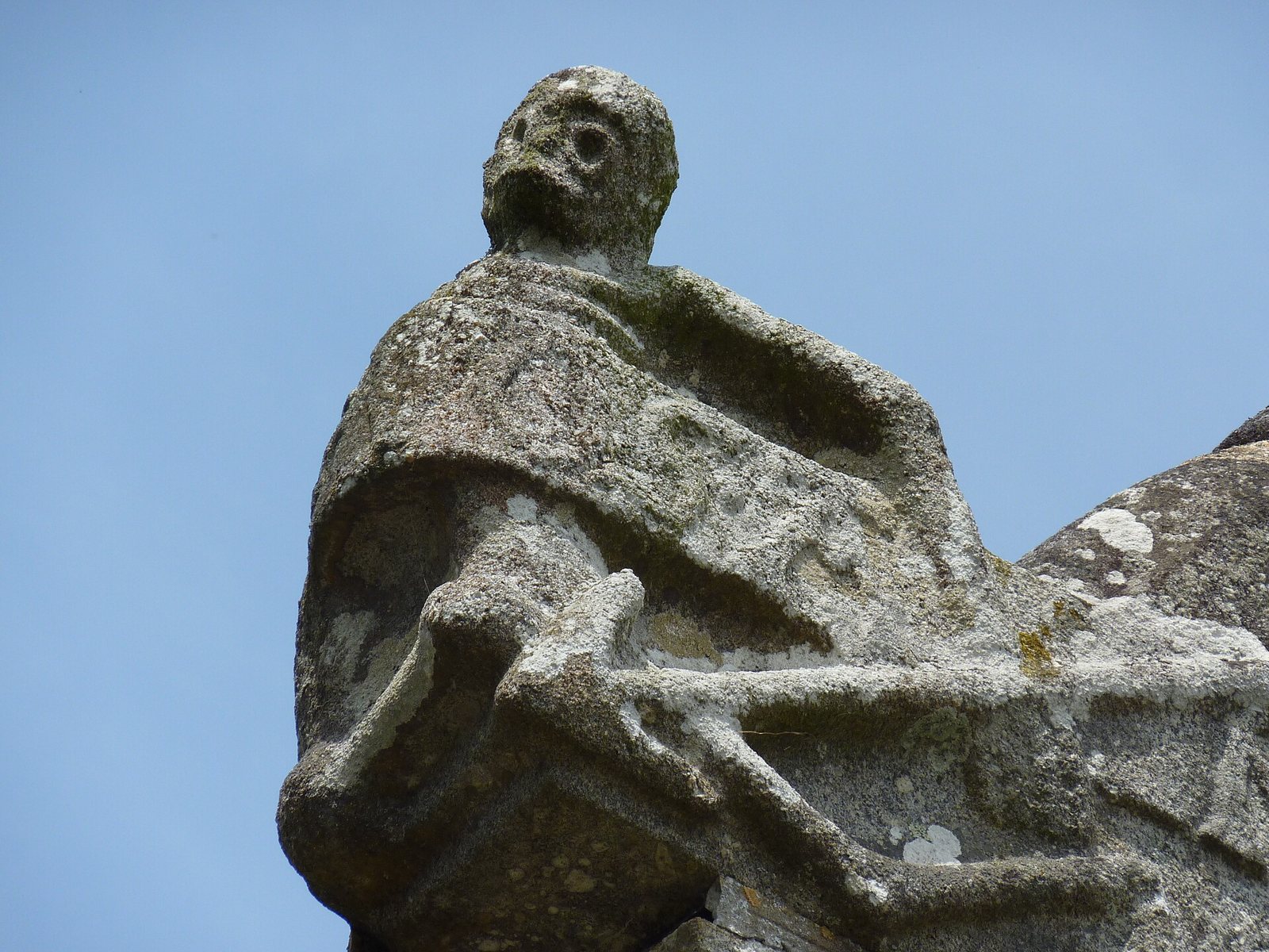 A weathered stone figure of the Ankou on the Brasparts ossuary, a hooded skeletal figure with a skull for a face, seen against a clear blue sky