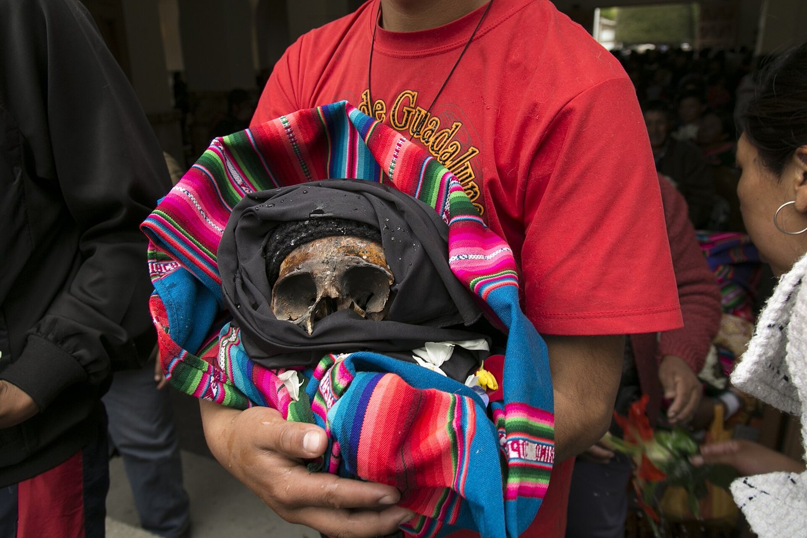 A person in a red T-shirt, face out of frame, holds a ñatita wrapped in black cloth and a brightly striped aguayo