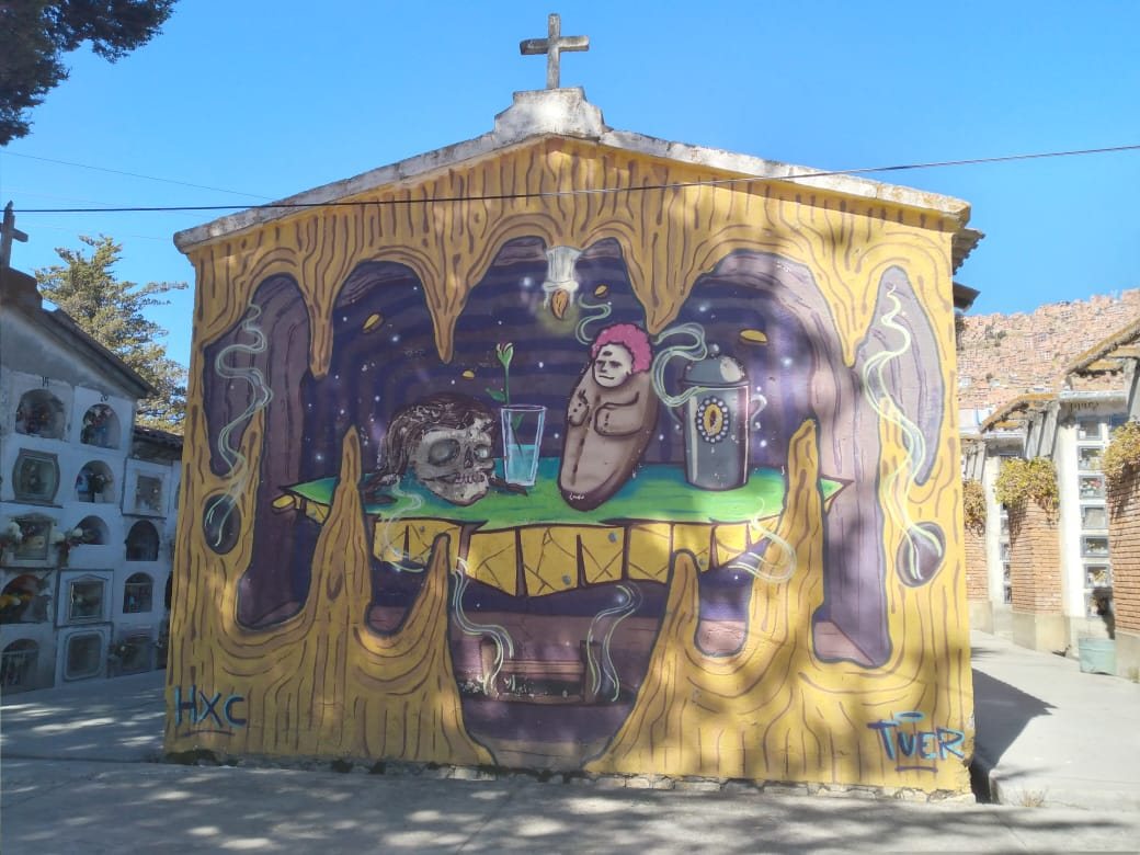 A small cemetery building painted with a mural of a flower-crowned skull and a small figure at a table, between walls of burial niches under a blue sky
