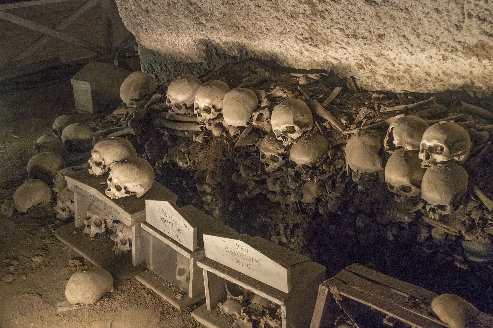Rows of skulls and long bones heaped along the base of a quarry wall at the Fontanelle cemetery, with small stone boxes and name plates set in front of them